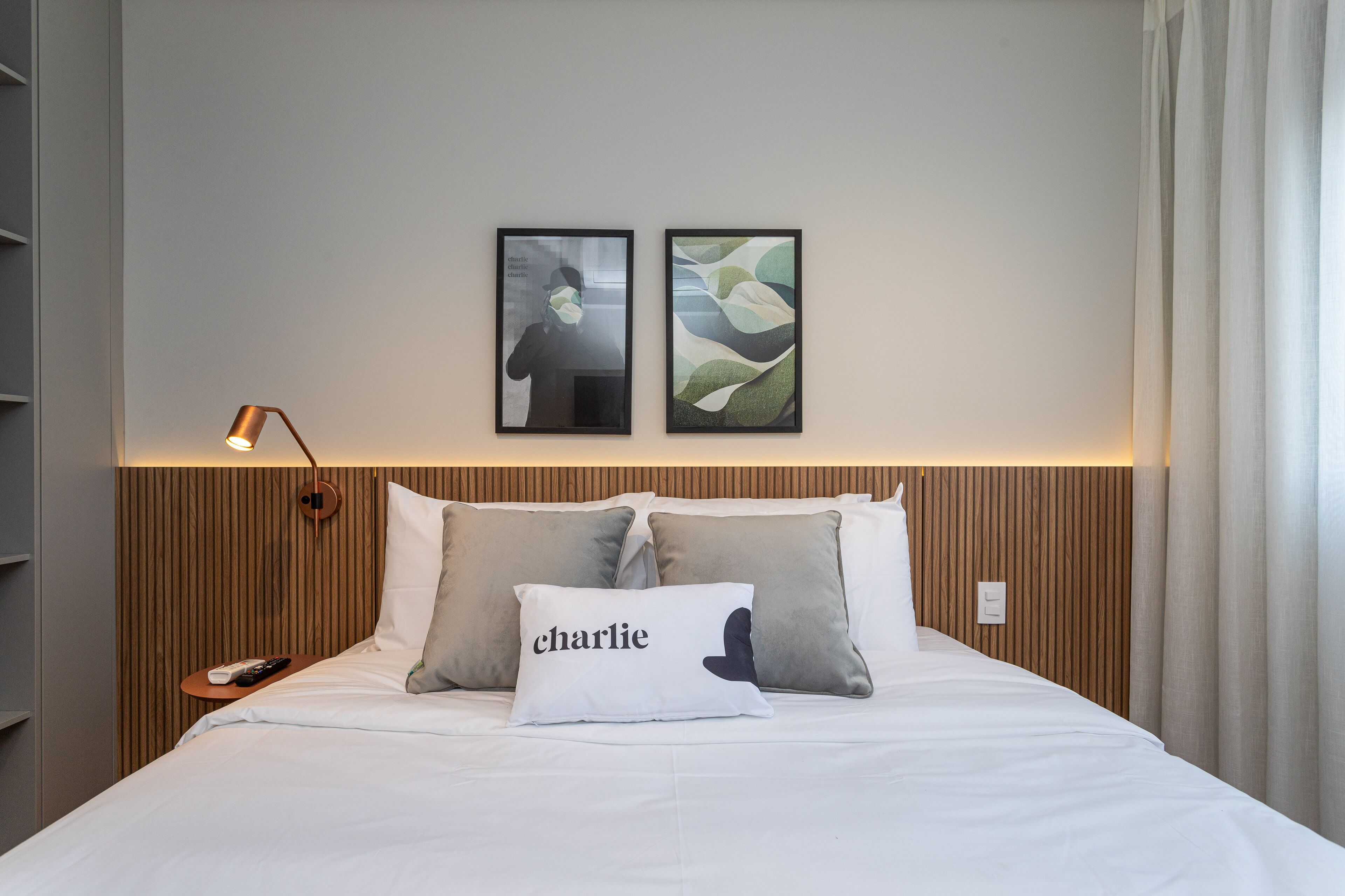 Ein ordentlich bezogenes Bett mit einem Kissen mit dem Aufdruck "charlie", ein Nachttisch mit Fernbedienung und zwei gerahmte Kunstwerke darüber.