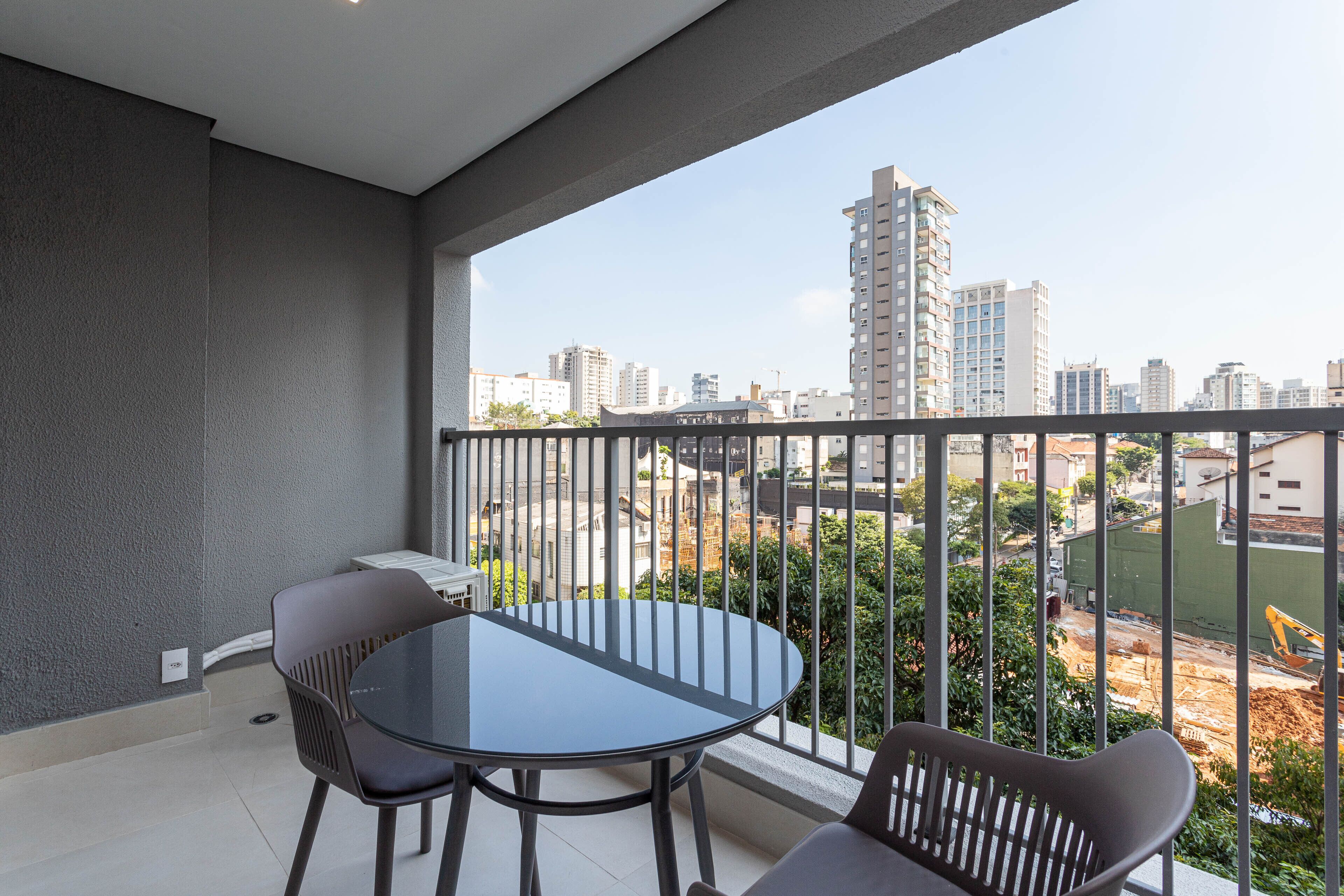Un balcon avec une table en verre et deux chaises, donnant sur un paysage urbain avec des immeubles et des chantiers de construction.