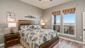 Une chambre avec un grand lit, deux tables de chevet avec des lampes, un ventilateur de plafond et une vue sur la plage grâce à de grandes fenêtres.