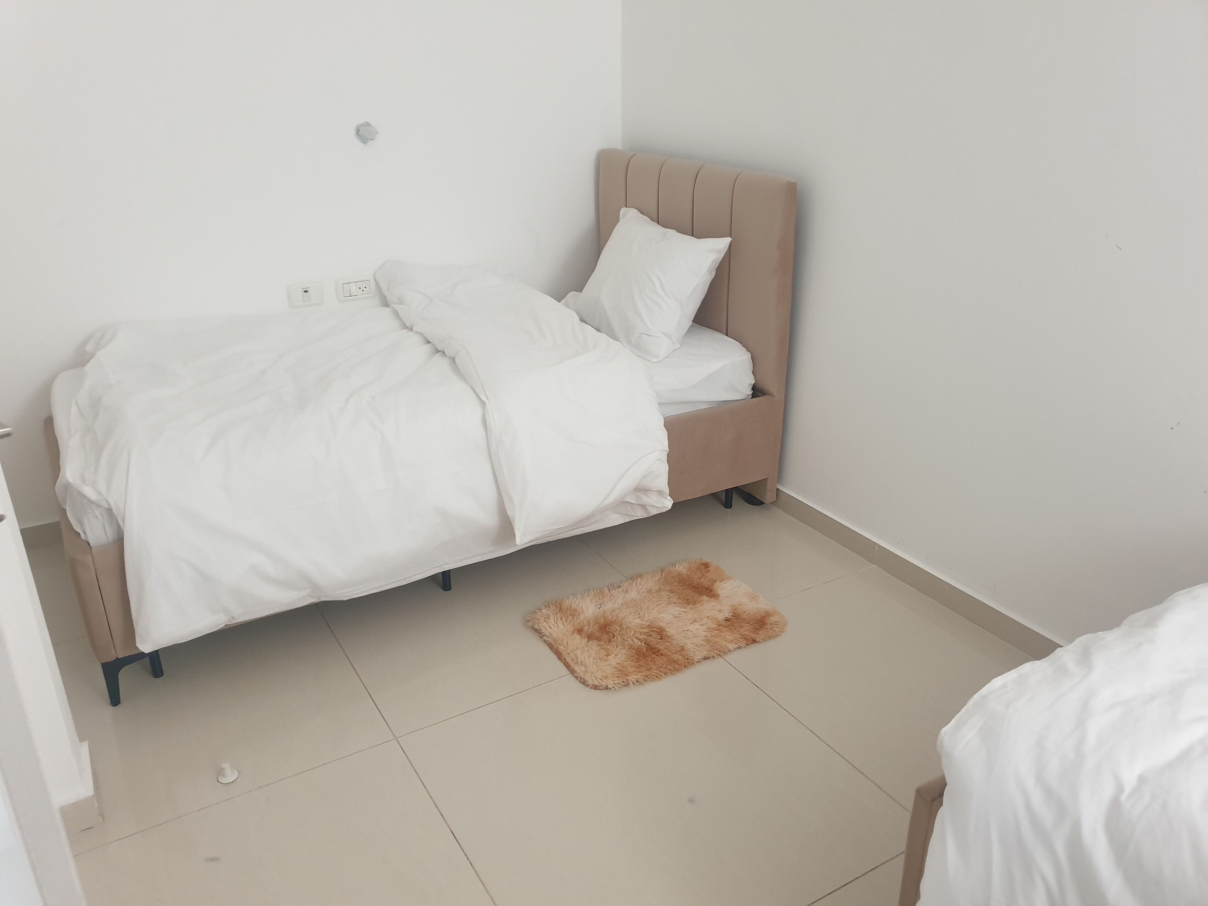 Un lit bien fait, avec une literie blanche, dans une pièce dont le sol est carrelé et où se trouve un petit tapis moelleux.