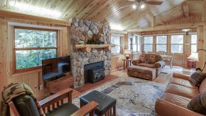 A cozy cabin interior with a stone fireplace, wooden furniture, and large windows offering a view of the forest. - Bay Lake Cabin by Historic Ruttger`s Resort (Deerwood)