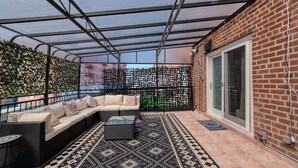 Una zona patio coperta con tetto in metallo nero, un tappeto a motivi bianchi e neri, un divano in vimini nero con cuscini e un tavolino rotondo.