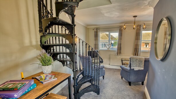 Un escalier orné de rampes noires, une table en bois avec des livres, une plante en pot et un fauteuil bleu dans une pièce donnant sur l’extérieur.