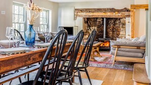 A dining area with a long wooden table, black chairs, and a blue vase with dried flowers. A stone fireplace with a fire is visible in the background. - Charming Downland Cottage - Pass the Keys (Chichester)