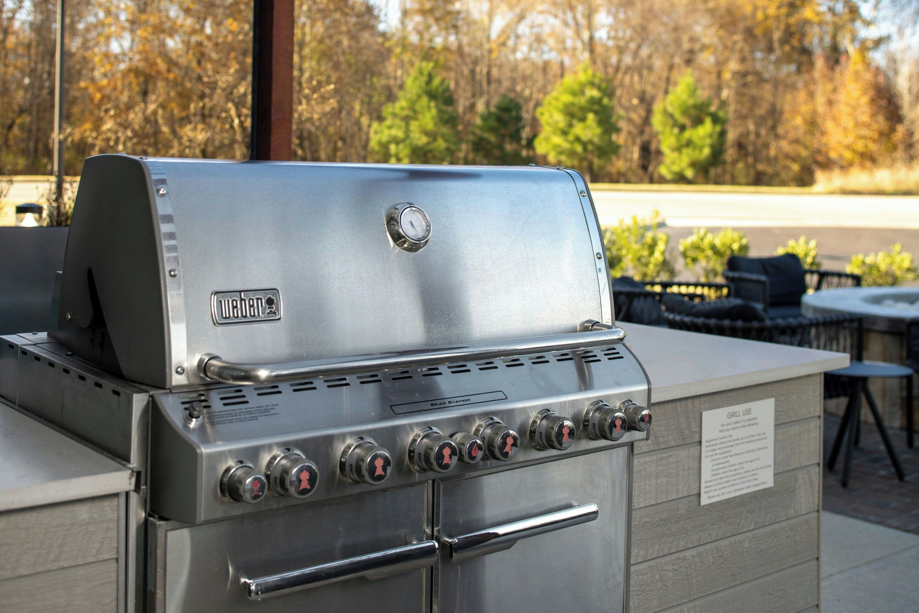 Ein Weber-Gasgrill aus Edelstahl mit mehreren Brennern und einer Seitentischablage, draußen gelegen mit Blick auf Bäume und einer Terrasse im Hintergrund.