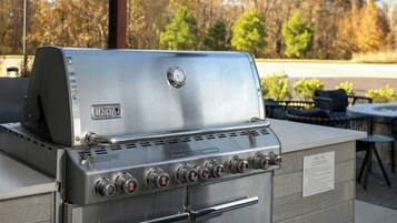 Ein Weber-Gasgrill aus Edelstahl mit mehreren Brennern und einer Seitentischablage, draußen gelegen mit Blick auf Bäume und einer Terrasse im Hintergrund.