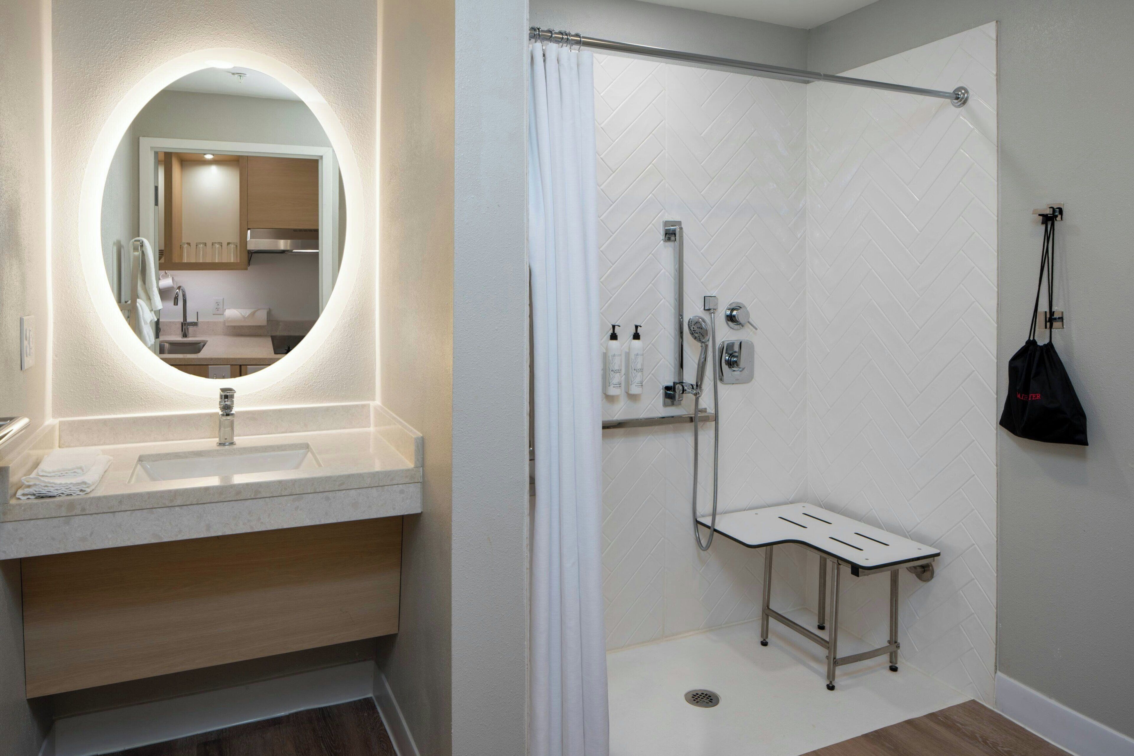 A modern bathroom with a round mirror, a sink, a shower with a handheld showerhead, and a small bench.