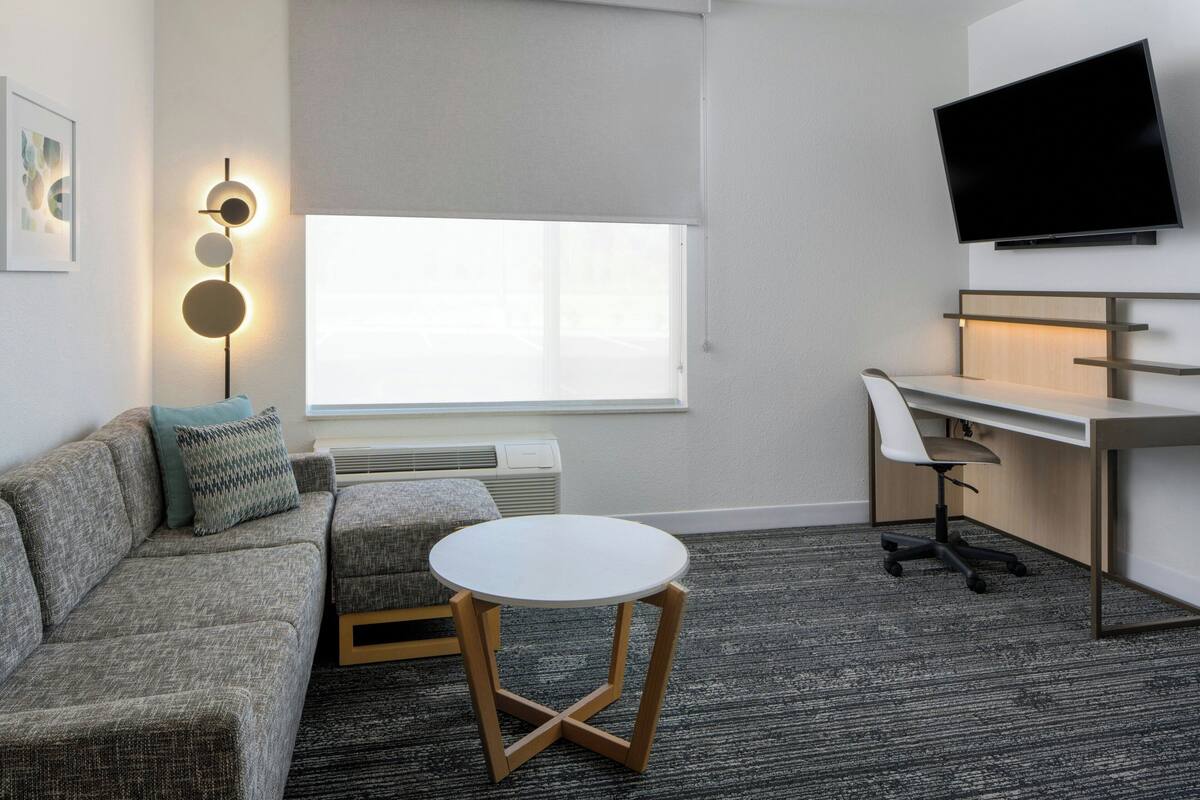Een moderne hotelkamer met een grijze bank, een klein rond tafeltje, een bureau met stoel, een flatscreen televisie aan de muur en een groot raam met uitzicht.