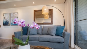 A modern living room with a grey sofa, a glass coffee table, and purple orchids. - Ocean View Condo in the Heart of Kihei! (Kihei)