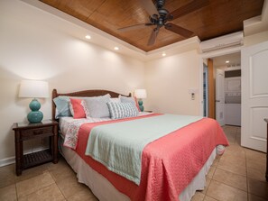 A bedroom with a wooden ceiling, a bed with multiple pillows, two bedside tables with lamps, and a door leading to a laundry area.