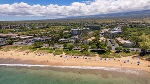 An aerial view of a coastal resort with multiple buildings, a sandy beach, and clear blue skies. - Ocean View Condo in the Heart of Kihei! (Kihei)