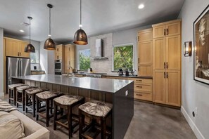 A modern kitchen with a central island, bar stools, wooden cabinets, pendant lighting, and a large window. - South Lamar Wellness Haven / Sauna / Hot Tub / BBQ (Austin)