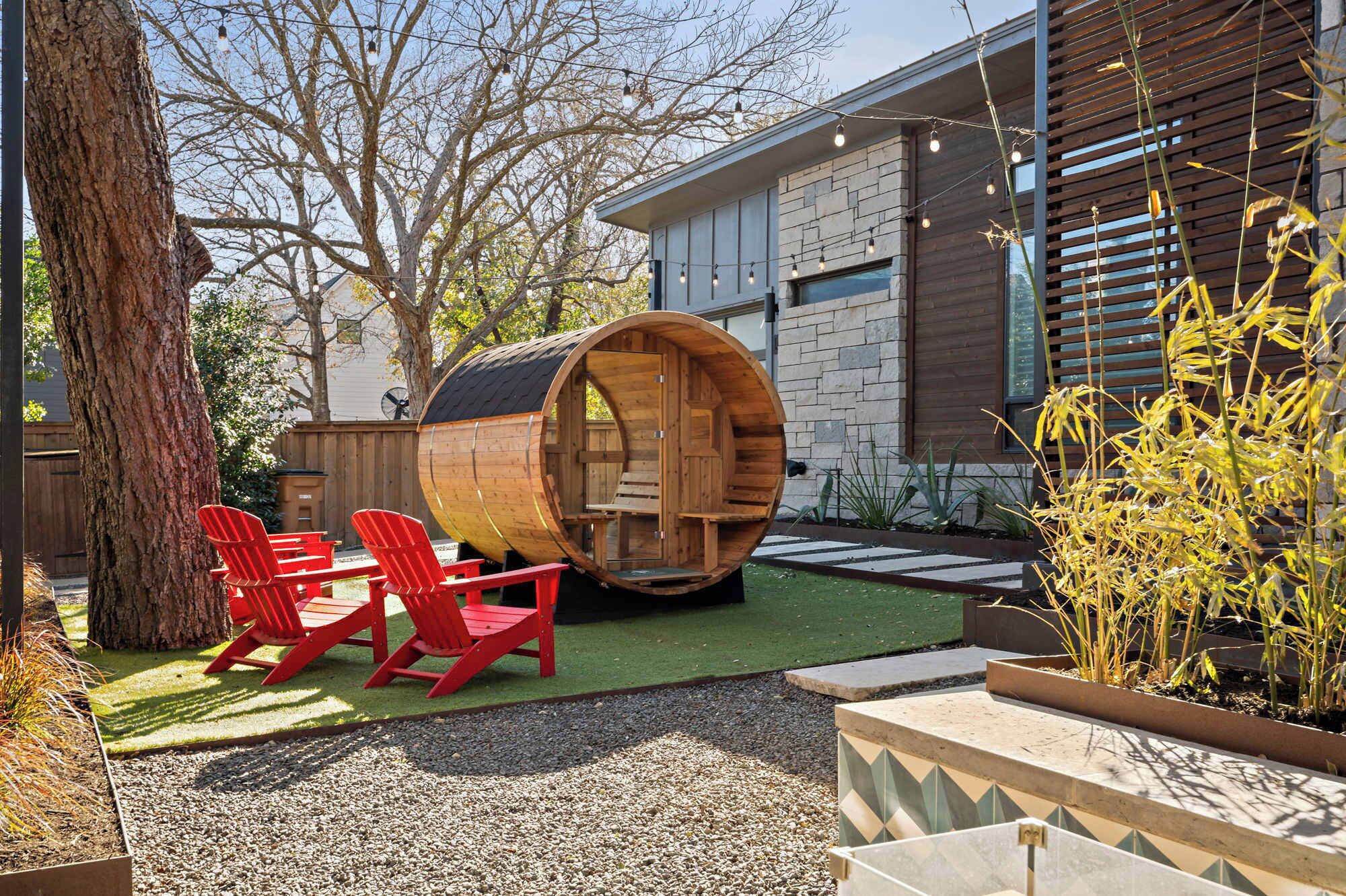 Un área de estar moderna al aire libre con un barril de madera, sillas rojas y un edificio de piedra al fondo.