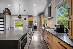 A modern kitchen with a central island, stainless steel appliances, wooden cabinets, pendant lighting, and a large window offering a view of greenery.