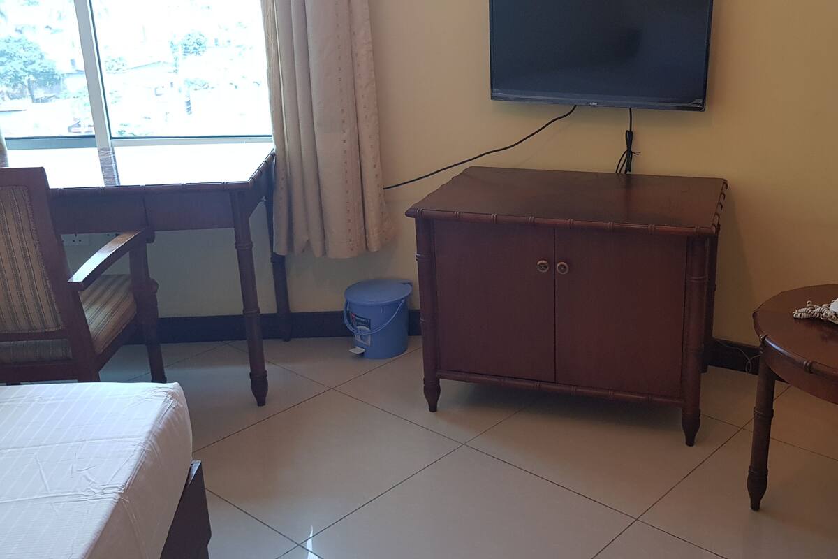 Une chambre d’hôtel avec un lit, une armoire en bois, une petite table avec un téléphone, un téléviseur fixé au mur et une fenêtre avec des rideaux.