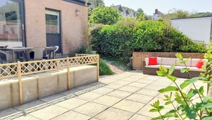 A patio area with a concrete floor, a wooden fence, a seating area with a sofa and cushions, and a building with a glass door and window. - 2 bedroom accommodation in Combe Martin (Combe Martin)