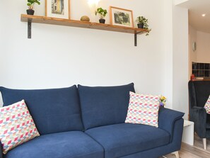 Een blauwe bank met gekleurde kussens, een houten plank met ingelijste foto’s en planten, en een klein witte tafeltje.