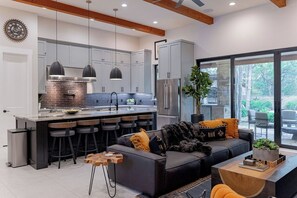 A modern kitchen with a central island, bar stools, and a dining area with a black sofa and orange chairs.