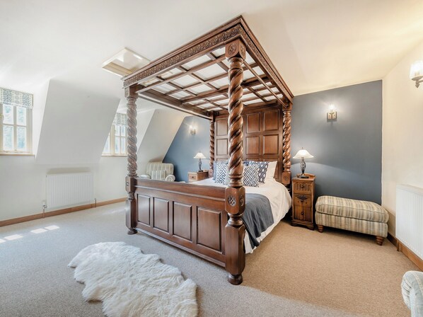 A four-poster bed with a wooden frame and a slatted canopy, a small bedside table with a lamp, a cushioned bench, and a fluffy white rug on the floor.