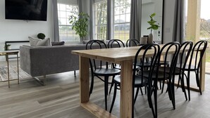 Uma sala de estar moderna com um sofá cinzento, uma mesa de jantar de madeira com cadeiras pretas e grandes janelas com cortinas.