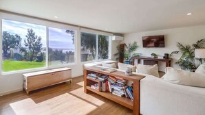 A modern living room with a large window, a bookshelf, a sofa, and a TV. - Ocean & Bay View Retreat | A/C & Private Yard (San Diego)