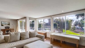 A spacious living room with a large window offering a view of the outdoors, a beige sofa, a bench, and a wooden coffee table. - Ocean & Bay View Retreat | A/C & Private Yard (San Diego)