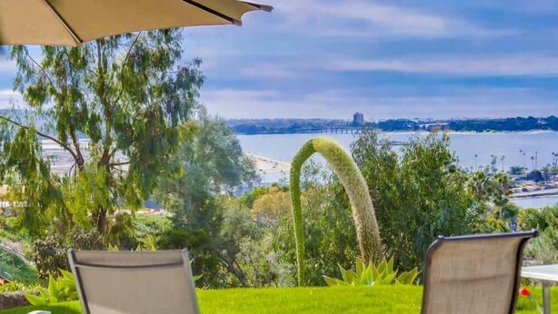 A patio with a view of a bridge and water, surrounded by greenery and a large umbrella. - Ocean & Bay View Retreat | A/C & Private Yard (San Diego)