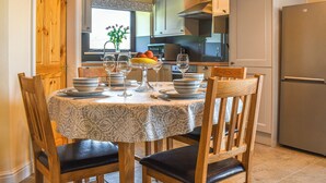 Un comedor con una mesa redonda dispuesta para cuatro personas, rodeada de sillas de madera con cojines negros. En la mesa hay un florero con flores, copas de vino y cuencos con frutas. La habitación tiene vigas de madera en el techo y se ve una cocina al fondo.