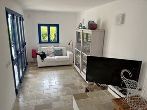 A living room with a white sofa, a television, a cabinet with glass doors, and a window with blue shutters.