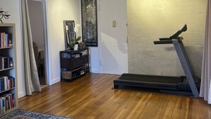 Een ruime woonkamer met een houten vloer, een loopband, een boekenkast en een groot vloerkleed.