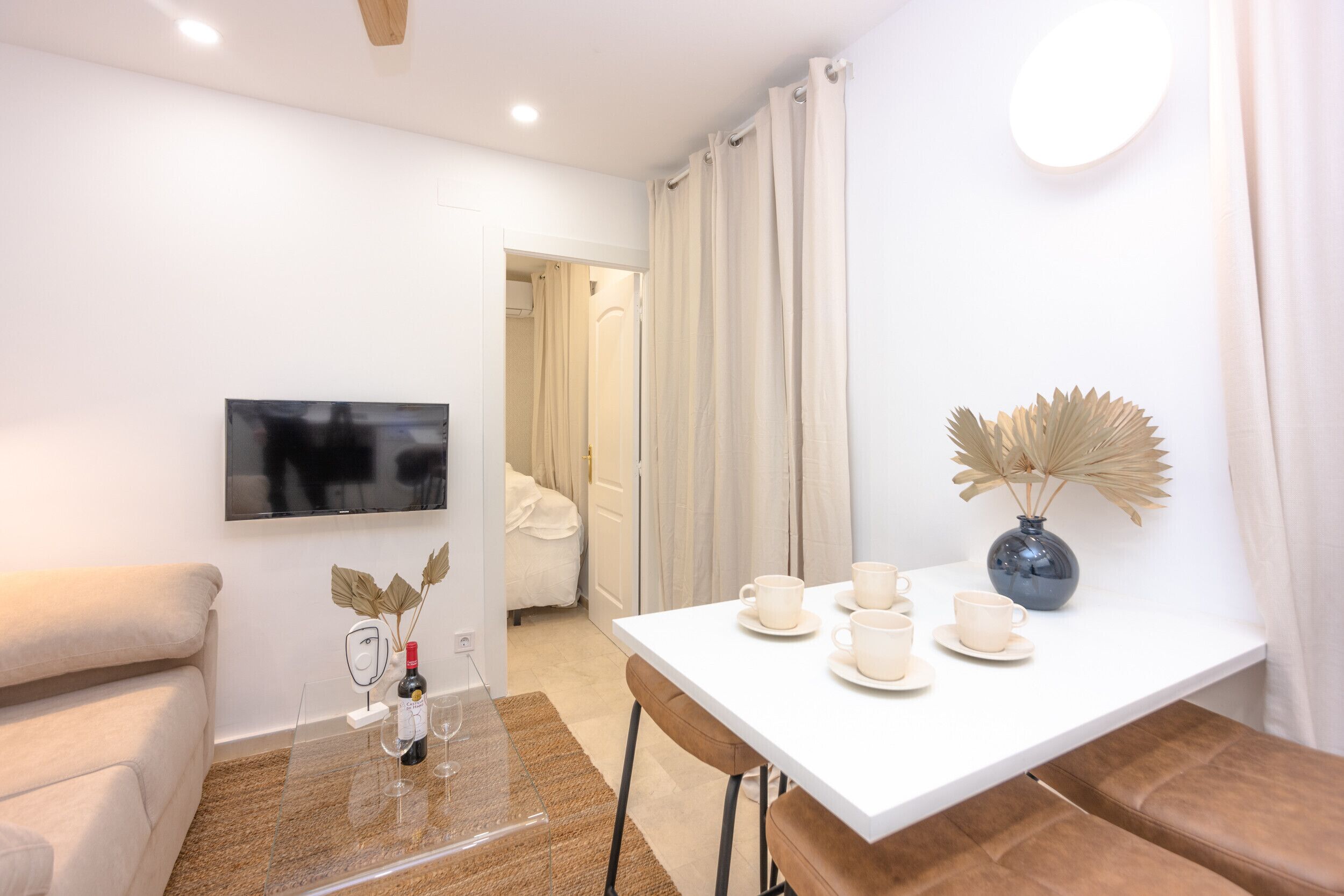 Une pièce moderne et bien éclairée, avec un canapé blanc, une table à manger et des chaises, un téléviseur fixé au mur et un vase contenant des fleurs séchées sur la table.
