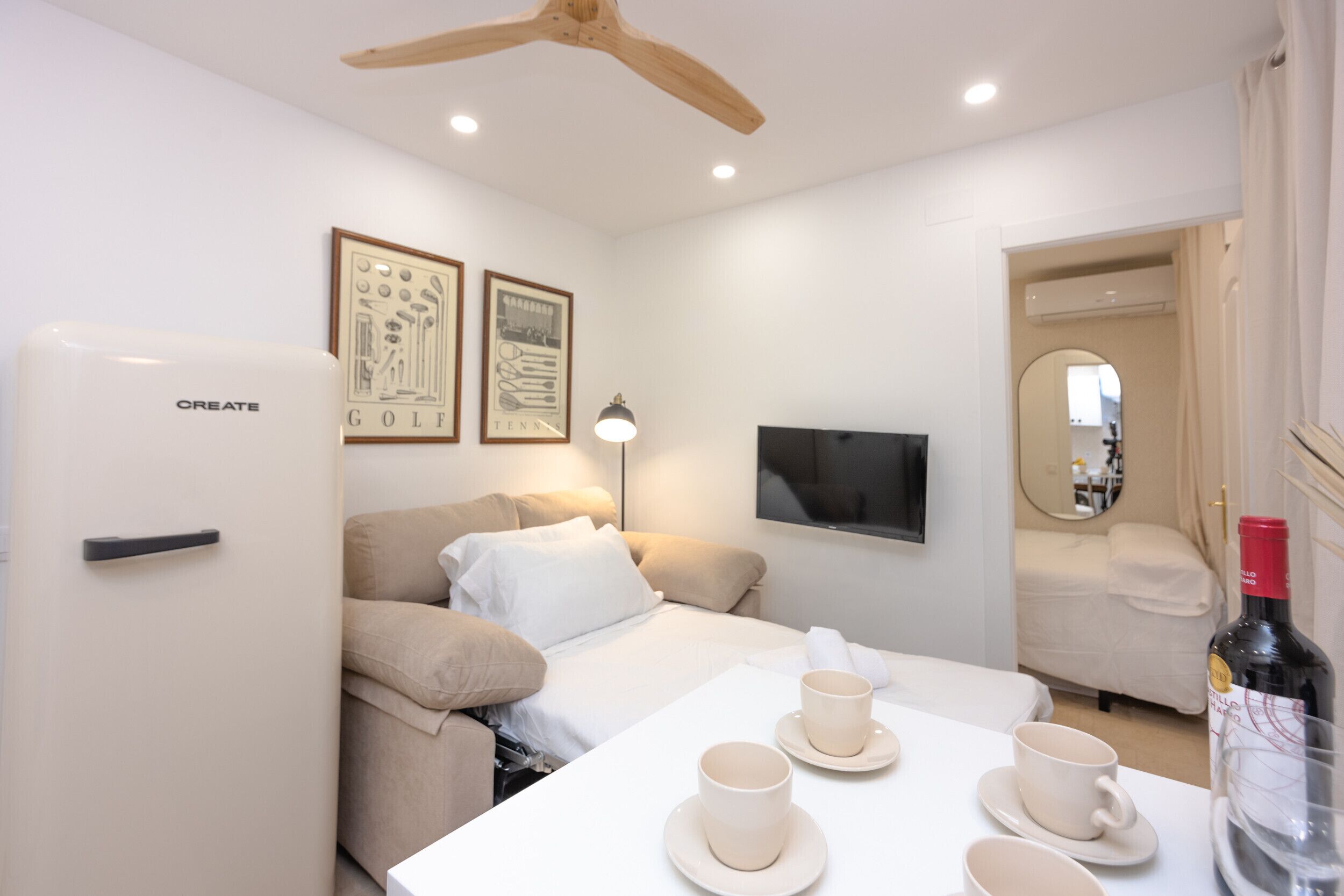 Habitación de hotel con cama, una mesa con tazas y platillos, refrigerador, ventilador de techo, cuadros en la pared y espejo.