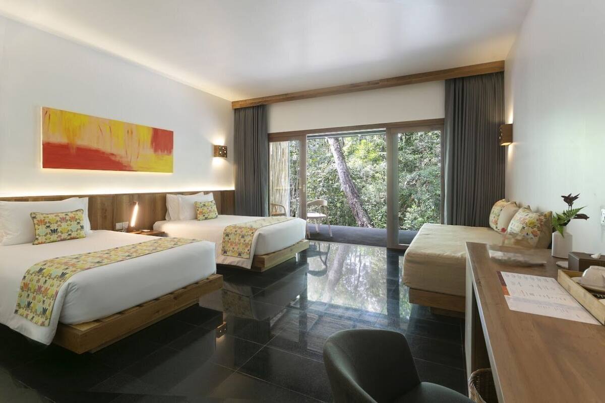 A hotel room with two beds, a desk, and a large window with a view of trees.