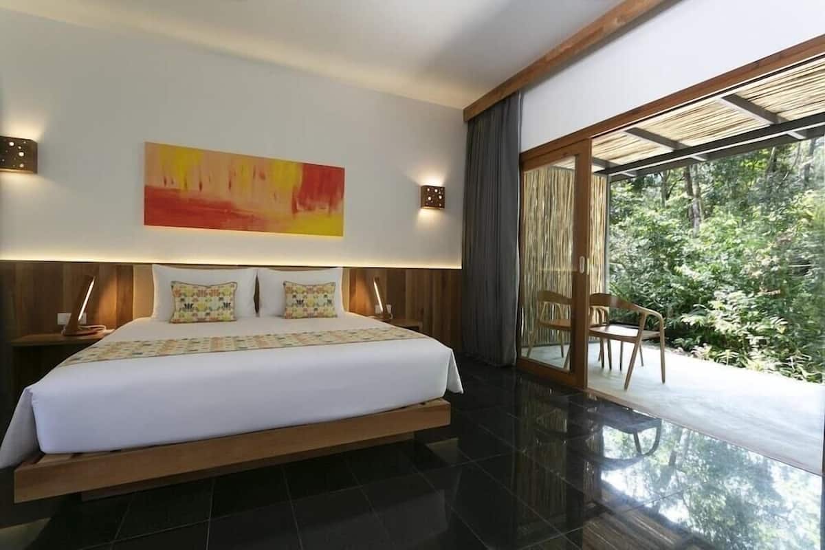 A modern bedroom with a large bed, a painting, and a view of greenery through a sliding glass door.
