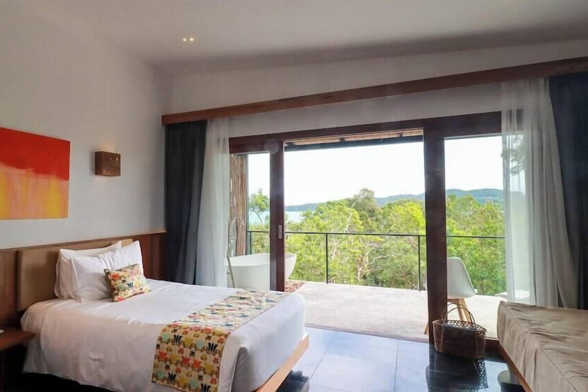 A hotel room with a large bed, a nightstand, a desk, a chair, and a balcony with a view of the landscape.