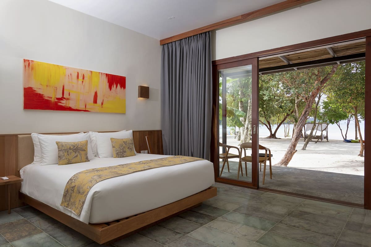 A modern hotel room with a large bed, a view of the beach, and a tropical landscape visible through the glass door.