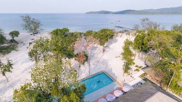 A pool surrounded by trees and a beach, with a view of the sea and boats.