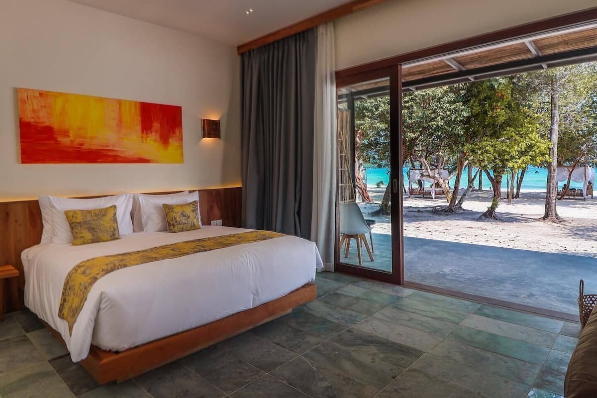 A bedroom with a large bed, a view of the beach, and a tropical tree outside.