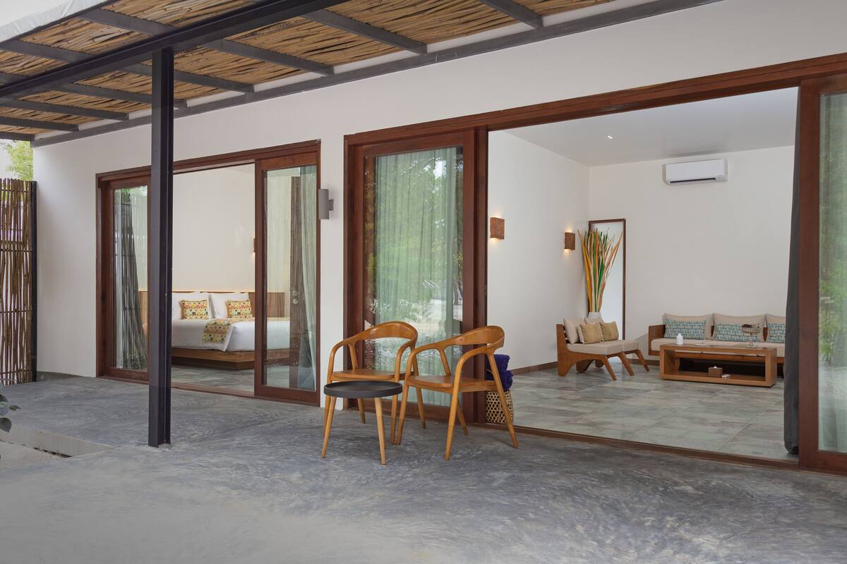 A spacious room with a large bed, two wooden chairs, a sofa, and a coffee table. There is a sliding glass door leading to an outdoor area.