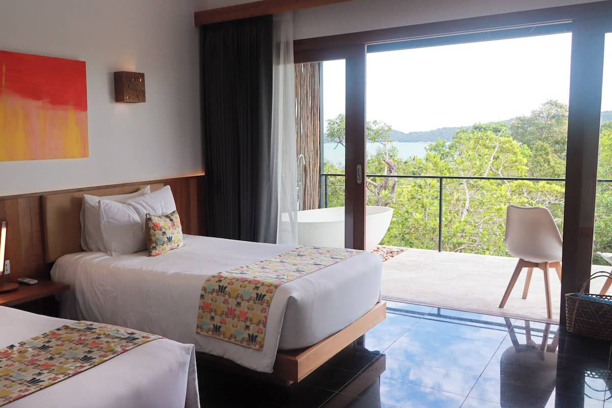 A hotel room with two beds, a large window with a view of greenery and hills, and a balcony with a chair.