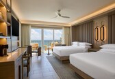 A hotel room with two beds, a desk, a chair, a TV, and a balcony with a view of the ocean.