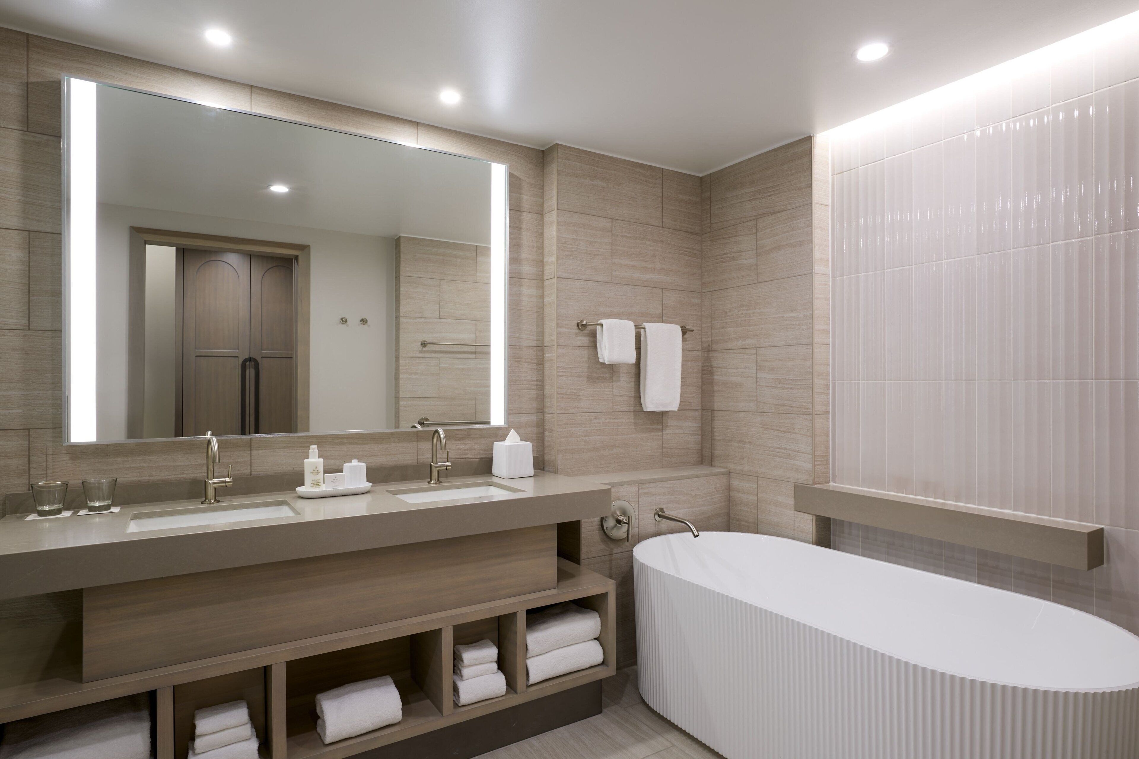 A modern bathroom with a large mirror, a double sink, a bathtub, and built-in shelves for towels.