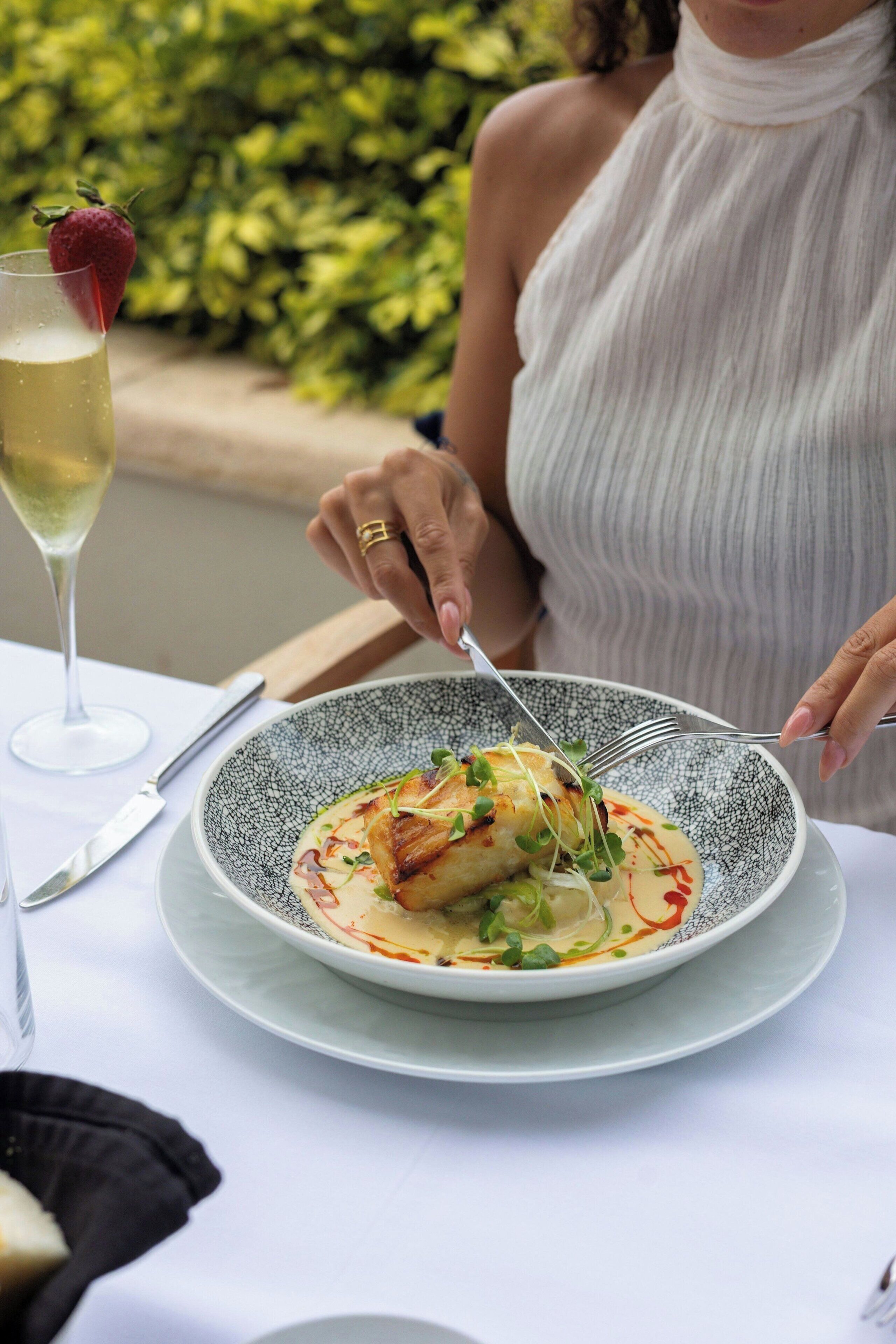A person dining at an outdoor table with a plate of food, a glass of white wine, and a fork.