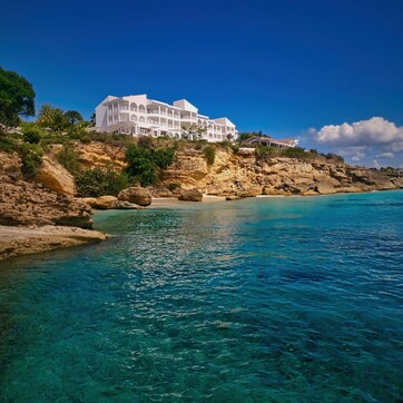 A coastal hotel with a white facade, situated on a rocky cliff overlooking turquoise waters.