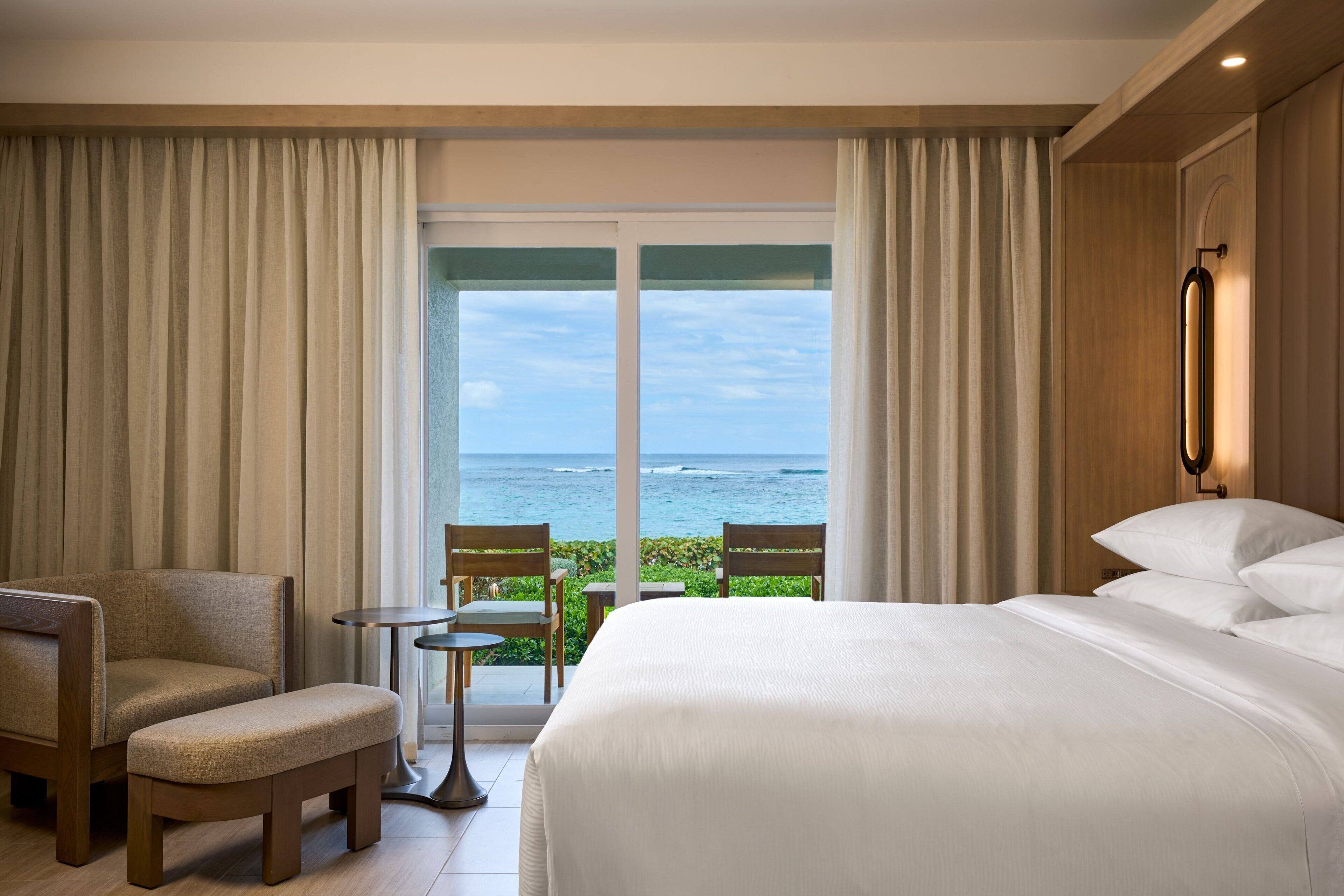 Une chambre d’hôtel avec un grand lit, deux fauteuils, une petite table et une vue sur l’océan par une grande fenêtre.