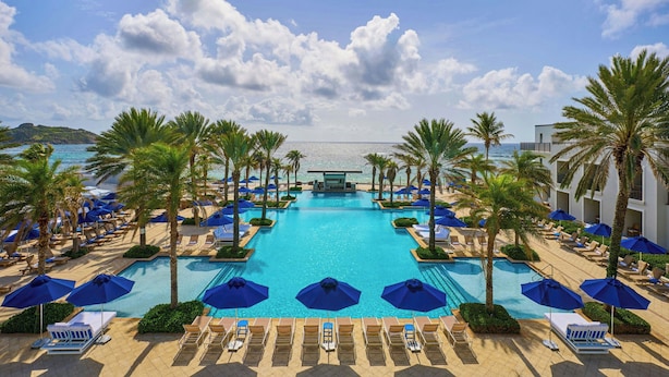 A pool area with lounge chairs and umbrellas, surrounded by palm trees, and a view of the sea.