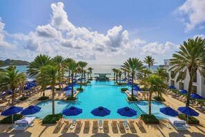 A pool area with lounge chairs and umbrellas, surrounded by palm trees, and a view of the sea.