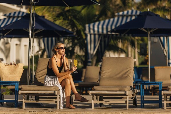 A person sitting on a lounge chair under a striped awning, holding a drink.