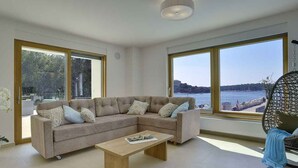 A modern living room with a large L-shaped sectional sofa, a wooden coffee table, and large windows offering a view of a body of water and distant land.