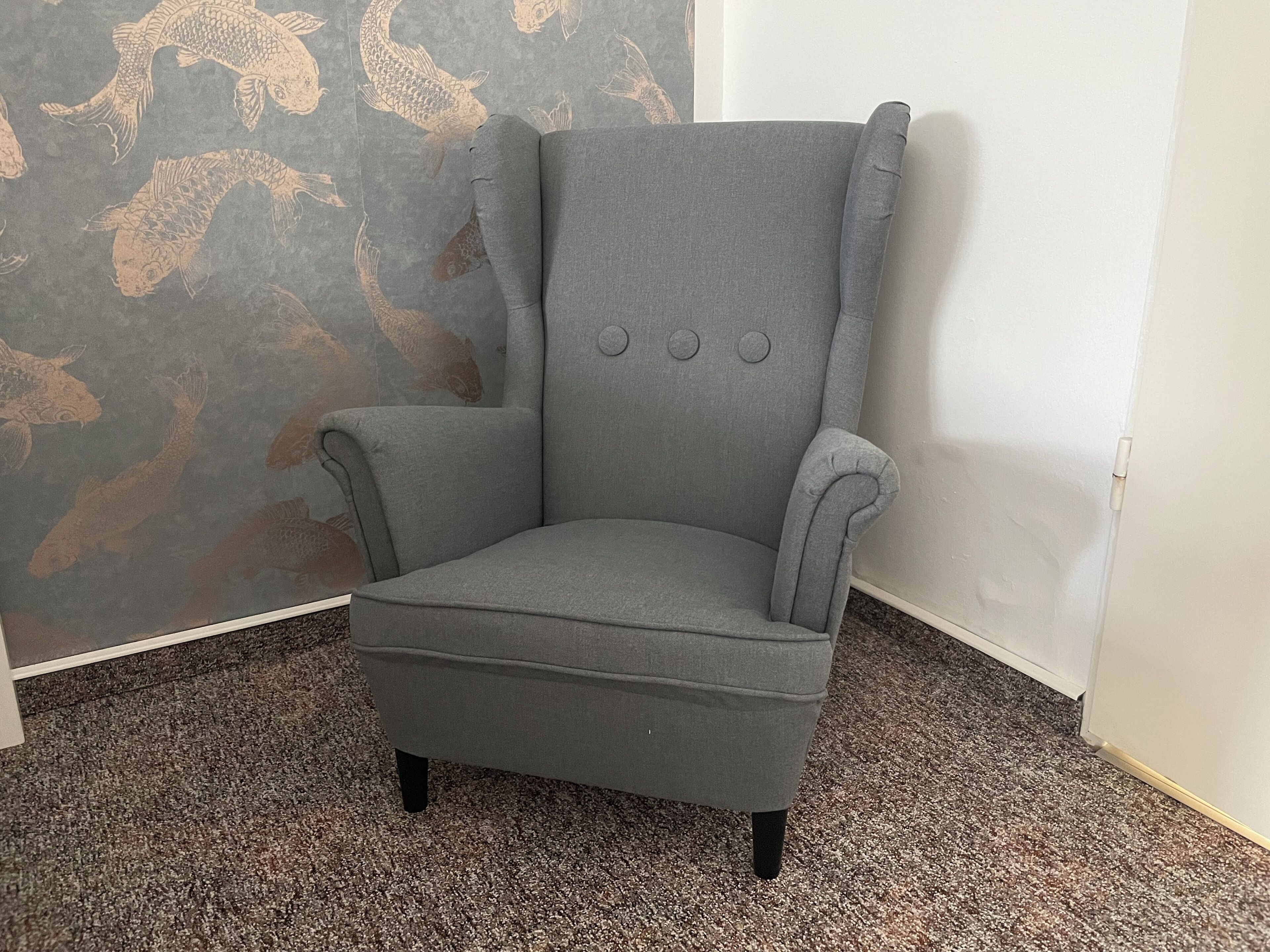 A grey armchair with a high back and armrests, positioned in a corner against a wall with a fish pattern.